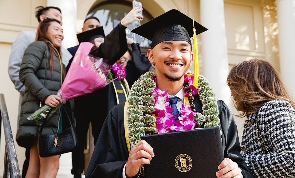 A young man wearing a black cap and gown at his graduation smiles at the camera.