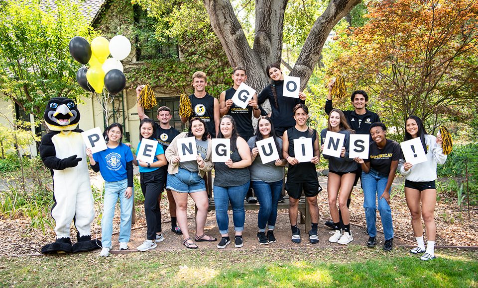 Group of students posing outdoors with Dominican University's penguin mascot, balloons, and cards spelling “GO PENGUINS.”