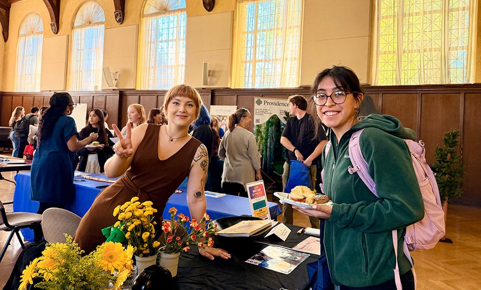 Two smiling women pose at an indoor job fair held in Guzman Hall on the Dominican campus ; one makes a peace sign while the other holds a plate of food.